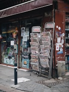 A street-side newsstand displays a variety of newspapers stacked in metal racks, positioned beside a shop window filled with various items. The storefront has a maroon awning and several posters, including some with images and text. The pavement is cobblestone, and a black-and-white striped pole is positioned in front.