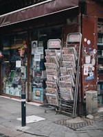 A street-side newsstand displays a variety of newspapers stacked in metal racks, positioned beside a shop window filled with various items. The storefront has a maroon awning and several posters, including some with images and text. The pavement is cobblestone, and a black-and-white striped pole is positioned in front.