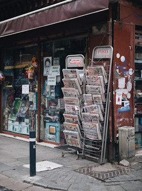 A street-side newsstand displays a variety of newspapers stacked in metal racks, positioned beside a shop window filled with various items. The storefront has a maroon awning and several posters, including some with images and text. The pavement is cobblestone, and a black-and-white striped pole is positioned in front.