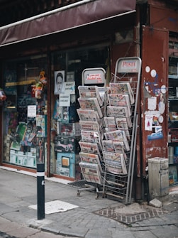 A street-side newsstand displays a variety of newspapers stacked in metal racks, positioned beside a shop window filled with various items. The storefront has a maroon awning and several posters, including some with images and text. The pavement is cobblestone, and a black-and-white striped pole is positioned in front.