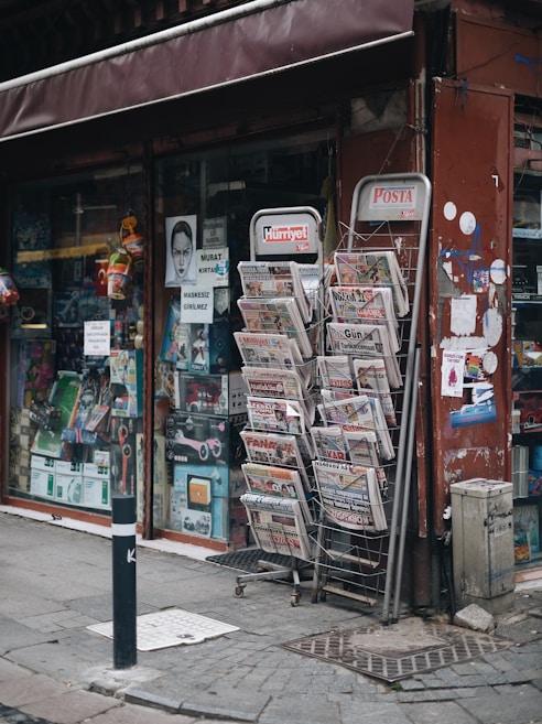 A street-side newsstand displays a variety of newspapers stacked in metal racks, positioned beside a shop window filled with various items. The storefront has a maroon awning and several posters, including some with images and text. The pavement is cobblestone, and a black-and-white striped pole is positioned in front.