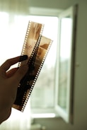 A hand is holding up two strips of photographic film negatives against a softly lit background with an open window. The film features multiple frames and is translucent, allowing light to pass through, revealing some details of the images captured on it.