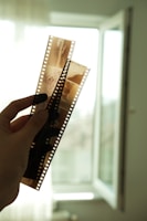 A hand is holding up two strips of photographic film negatives against a softly lit background with an open window. The film features multiple frames and is translucent, allowing light to pass through, revealing some details of the images captured on it.