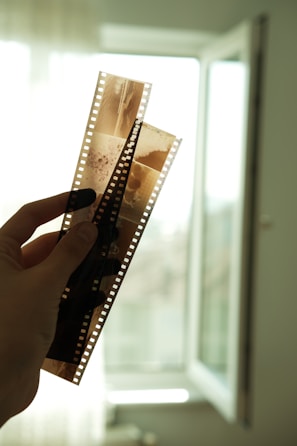 A hand is holding up two strips of photographic film negatives against a softly lit background with an open window. The film features multiple frames and is translucent, allowing light to pass through, revealing some details of the images captured on it.