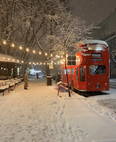 A cozy London street covered in winter snow with iconic red buses passing by.