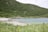 A serene coastal landscape featuring a sandy beach that curves along a turquoise bay. Lush green grass and wildflowers are in the foreground, transitioning into dense tree-covered hills. A small, red-roofed cabin is nestled among the greenery, providing a sense of isolation and tranquility. The sky appears overcast, contributing to the peaceful atmosphere of the setting.
