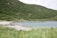 A serene coastal landscape featuring a sandy beach that curves along a turquoise bay. Lush green grass and wildflowers are in the foreground, transitioning into dense tree-covered hills. A small, red-roofed cabin is nestled among the greenery, providing a sense of isolation and tranquility. The sky appears overcast, contributing to the peaceful atmosphere of the setting.