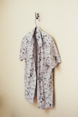 A light-colored shirt with black printed patterns hangs on a blue hanger. The shirt features tropical designs and text, and it is slightly unbuttoned. It is displayed against a plain beige wall using a single metal hook.