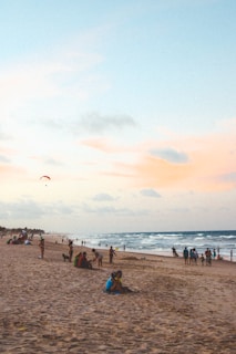 A sandy beach with groups of people enjoying the oceanfront, some sitting and others walking along the shore. The waves gently lap against the beach under a sky with soft pastel hues of blue, pink, and orange. A parasailor is visible in the distance, adding a sense of adventure to the tranquil scene.