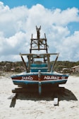 A traditional fishing boat with the words 'Japão Aquiraz CE' painted on it is resting on a sandy beach, supported by logs. The boat is made of wood, painted in blue and red, with a rustic, worn appearance. In the background, there are some small houses and lush green trees under a bright blue sky with fluffy white clouds.