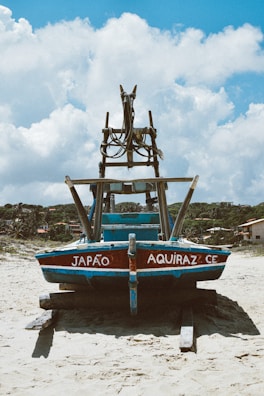 A traditional fishing boat with the words 'Japão Aquiraz CE' painted on it is resting on a sandy beach, supported by logs. The boat is made of wood, painted in blue and red, with a rustic, worn appearance. In the background, there are some small houses and lush green trees under a bright blue sky with fluffy white clouds.