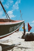 A small boat rests on a sandy beach with the words 'REI DO MAR' painted on its side. The background features several colorful sails and blue sky with some clouds. The scene suggests a coastal or beach environment, with visible details of sand and scattered vegetation.