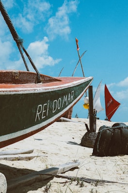 A small boat rests on a sandy beach with the words 'REI DO MAR' painted on its side. The background features several colorful sails and blue sky with some clouds. The scene suggests a coastal or beach environment, with visible details of sand and scattered vegetation.