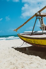 A traditional fishing boat rests on a sandy beach with the ocean waves gently rolling in the background under a clear blue sky. The boat is painted yellow with the word 'HOLANDA I' visible on its side. The beach appears serene and empty.