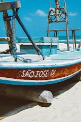 A wooden fishing boat with the name SÃO JOSE IX painted in white is resting on a sandy beach. The boat is primarily blue with a brown and white trim. Thick blue ropes are coiled around wooden posts on the deck, and the ocean is visible in the background under a clear blue sky.