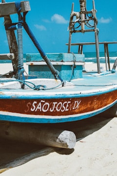 A wooden fishing boat with the name SÃO JOSE IX painted in white is resting on a sandy beach. The boat is primarily blue with a brown and white trim. Thick blue ropes are coiled around wooden posts on the deck, and the ocean is visible in the background under a clear blue sky.