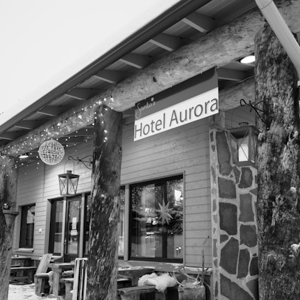 An exterior view of a building labeled 'Hotel Aurora' with rustic wooden elements and decor. The building features a stone and wood facade, with a sign hanging under the eaves. String lights and lanterns add to the decor, and there is snow on the ground and seating furniture outside the entrance.