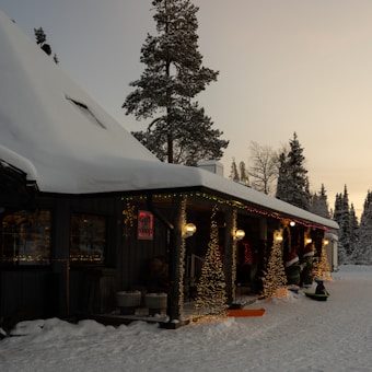 A cozy, snow-covered cabin surrounded by trees in a winter landscape. The cabin has a decorated porch with string lights and festive decorations. A sign indicates it's a gift shop. The sun is setting, casting a warm glow over the snowy scene.
