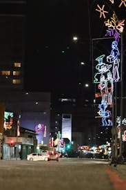 A street at night is adorned with vibrant neon lights. Various colorful neon figures and shapes hang along the roadside, including human-like figures conveying festive imagery. The street is dimly lit, with a few cars visible and buildings lined with additional decorations. The scene has an urban yet festive atmosphere.