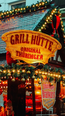 A festive wooden stall sign with 'Grill Hütte Original Thüringer' is adorned with Christmas decorations, including garlands and warm lights. The stall also advertises 'Thüringer Bratwurst' and features a nutcracker decoration.