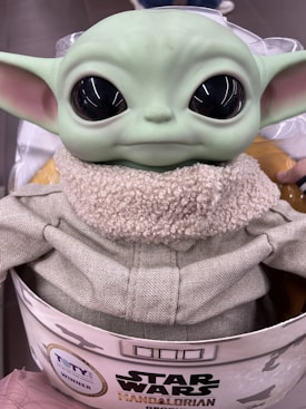 A figurine of a small green character with large black eyes and prominent ears, dressed in a cozy textured outfit. The packaging features branding related to Star Wars and The Mandalorian, and there is a label indicating a toy award win.