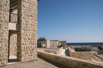 Stone buildings with rustic architecture are set against a clear blue sky and an expansive view of the ocean. The foreground features a detailed stone wall, with a glimpse of a wooden staircase inside an archway. Vegetation can be seen scattered along the pathways, adding a natural touch to the setting.