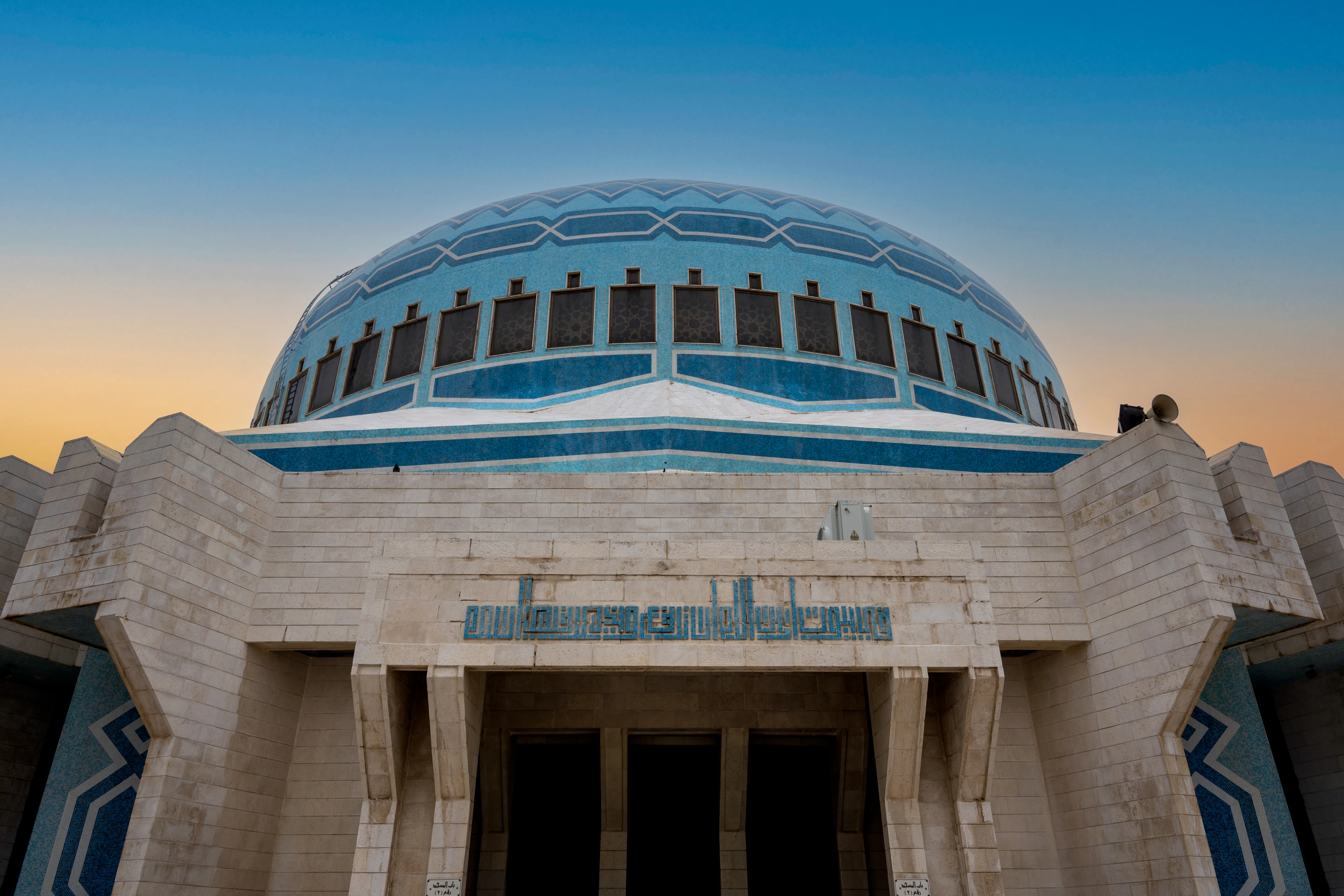 The blue and white mosaic dome of the King Abdullah I Mosque in Amman rising above its pale limestone portal, seen from below against a sky graduating from deep blue to gold at sunset.