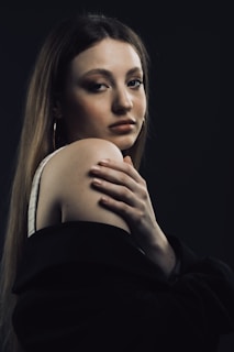 A woman gazes thoughtfully with her hand gently resting on her shoulder. She is dressed in a dark jacket, partially off her shoulder. Her expression is calm and introspective, and her long hair falls straight down her back. The background is dark, creating a moody atmosphere.