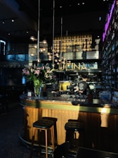 A warmly lit interior of a bar or restaurant, featuring a wooden counter with a marble top. Tall stools are placed at the bar, and a person is working behind the counter. Hanging spherical lights and a bouquet of pink lilies add to the decor. The word 'CUCINA' is displayed prominently above the bar area. Shelves filled with bottles line the walls, and a neon sign adds a modern touch.