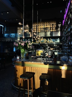 A warmly lit interior of a bar or restaurant, featuring a wooden counter with a marble top. Tall stools are placed at the bar, and a person is working behind the counter. Hanging spherical lights and a bouquet of pink lilies add to the decor. The word 'CUCINA' is displayed prominently above the bar area. Shelves filled with bottles line the walls, and a neon sign adds a modern touch.