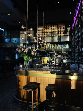A warmly lit interior of a bar or restaurant, featuring a wooden counter with a marble top. Tall stools are placed at the bar, and a person is working behind the counter. Hanging spherical lights and a bouquet of pink lilies add to the decor. The word 'CUCINA' is displayed prominently above the bar area. Shelves filled with bottles line the walls, and a neon sign adds a modern touch.