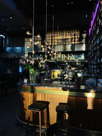 A warmly lit interior of a bar or restaurant, featuring a wooden counter with a marble top. Tall stools are placed at the bar, and a person is working behind the counter. Hanging spherical lights and a bouquet of pink lilies add to the decor. The word 'CUCINA' is displayed prominently above the bar area. Shelves filled with bottles line the walls, and a neon sign adds a modern touch.