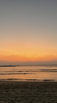 The image depicts a serene beach at sunset, with gentle waves lapping against the shore. The sky is a gradient of warm colors, transitioning from orange near the horizon to darker blues higher up. The calm water reflects the colors of the sky, creating a peaceful and tranquil scene.