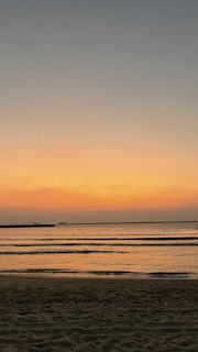 The image depicts a serene beach at sunset, with gentle waves lapping against the shore. The sky is a gradient of warm colors, transitioning from orange near the horizon to darker blues higher up. The calm water reflects the colors of the sky, creating a peaceful and tranquil scene.
