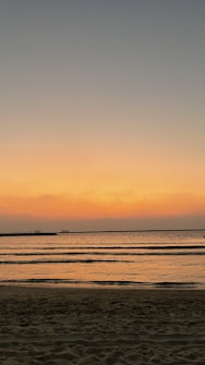 The image depicts a serene beach at sunset, with gentle waves lapping against the shore. The sky is a gradient of warm colors, transitioning from orange near the horizon to darker blues higher up. The calm water reflects the colors of the sky, creating a peaceful and tranquil scene.