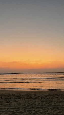 The image depicts a serene beach at sunset, with gentle waves lapping against the shore. The sky is a gradient of warm colors, transitioning from orange near the horizon to darker blues higher up. The calm water reflects the colors of the sky, creating a peaceful and tranquil scene.