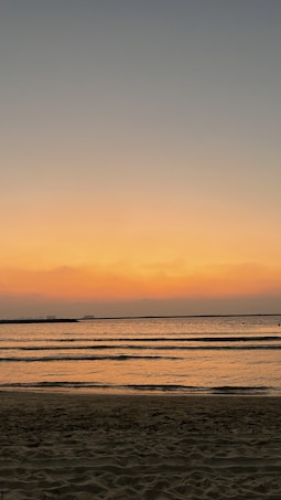 The image depicts a serene beach at sunset, with gentle waves lapping against the shore. The sky is a gradient of warm colors, transitioning from orange near the horizon to darker blues higher up. The calm water reflects the colors of the sky, creating a peaceful and tranquil scene.