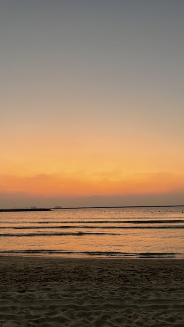 The image depicts a serene beach at sunset, with gentle waves lapping against the shore. The sky is a gradient of warm colors, transitioning from orange near the horizon to darker blues higher up. The calm water reflects the colors of the sky, creating a peaceful and tranquil scene.