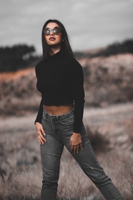 A woman stands outdoors with a confident posture, wearing a black long-sleeve top and gray jeans. She accessorizes with large sunglasses and has long dark hair. The background consists of blurred natural scenery, suggesting an overcast day.