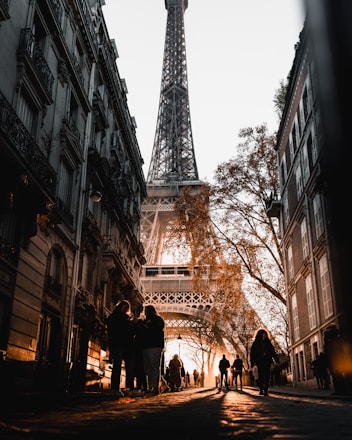 A street scene in Paris with the Eiffel Tower towering in the background, framed by buildings on both sides. The warm glow of the sunset casts long shadows and illuminates the urban setting. People are walking along the cobblestone path, and autumn trees add a touch of nature to the cityscape.