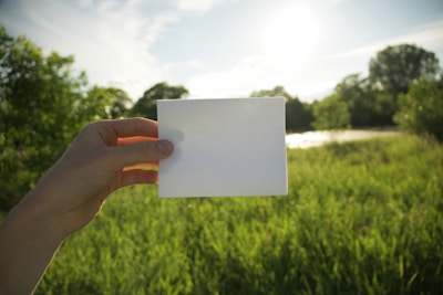 A hand holding a blank white piece of paper against a background of lush green grass and trees under a bright sunlit sky.
