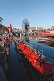 A vibrant amusement park scene featuring a large Ferris wheel with a smiling cartoon face in the center. Bright red roller coaster cars filled with people are on a track beside a body of water, reflecting the clear blue sky. The area is bustling with activity and surrounded by palm trees and buildings.