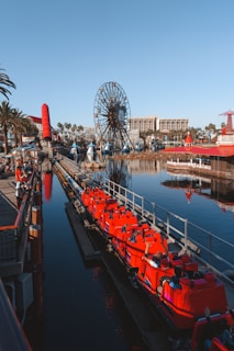A vibrant amusement park scene featuring a large Ferris wheel with a smiling cartoon face in the center. Bright red roller coaster cars filled with people are on a track beside a body of water, reflecting the clear blue sky. The area is bustling with activity and surrounded by palm trees and buildings.