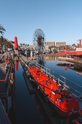 A vibrant amusement park scene featuring a large Ferris wheel with a smiling cartoon face in the center. Bright red roller coaster cars filled with people are on a track beside a body of water, reflecting the clear blue sky. The area is bustling with activity and surrounded by palm trees and buildings.
