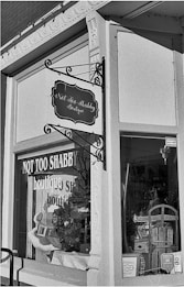 A black and white photo of a storefront with a sign that reads 'Not Too Shabby Boutique'. The window displays various items including a wreath, decorative boxes, and a chair. The store has ornate architectural details around the window frame and a classic hanging sign.