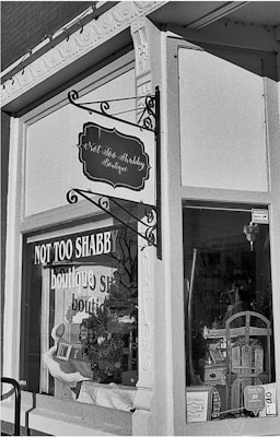A black and white photo of a storefront with a sign that reads 'Not Too Shabby Boutique'. The window displays various items including a wreath, decorative boxes, and a chair. The store has ornate architectural details around the window frame and a classic hanging sign.