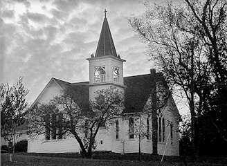 A historic church building with a prominent bell tower and cross, surrounded by trees. The architecture is traditional with pointed arch windows and wood-panelled exterior, set under a cloudy sky.