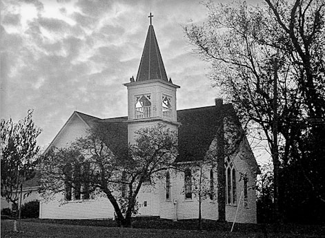 A historic church building with a prominent bell tower and cross, surrounded by trees. The architecture is traditional with pointed arch windows and wood-panelled exterior, set under a cloudy sky.