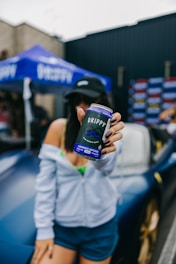 A person holds a can labeled 'DRIPPY' with the text 'cannabis infused' on it. The person is standing in front of a car, wearing a cap and casual attire. A blue tent with a brand name is in the background.