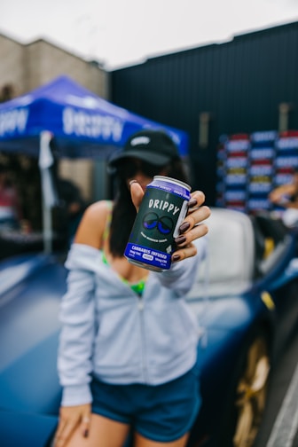 A person holds a can labeled 'DRIPPY' with the text 'cannabis infused' on it. The person is standing in front of a car, wearing a cap and casual attire. A blue tent with a brand name is in the background.